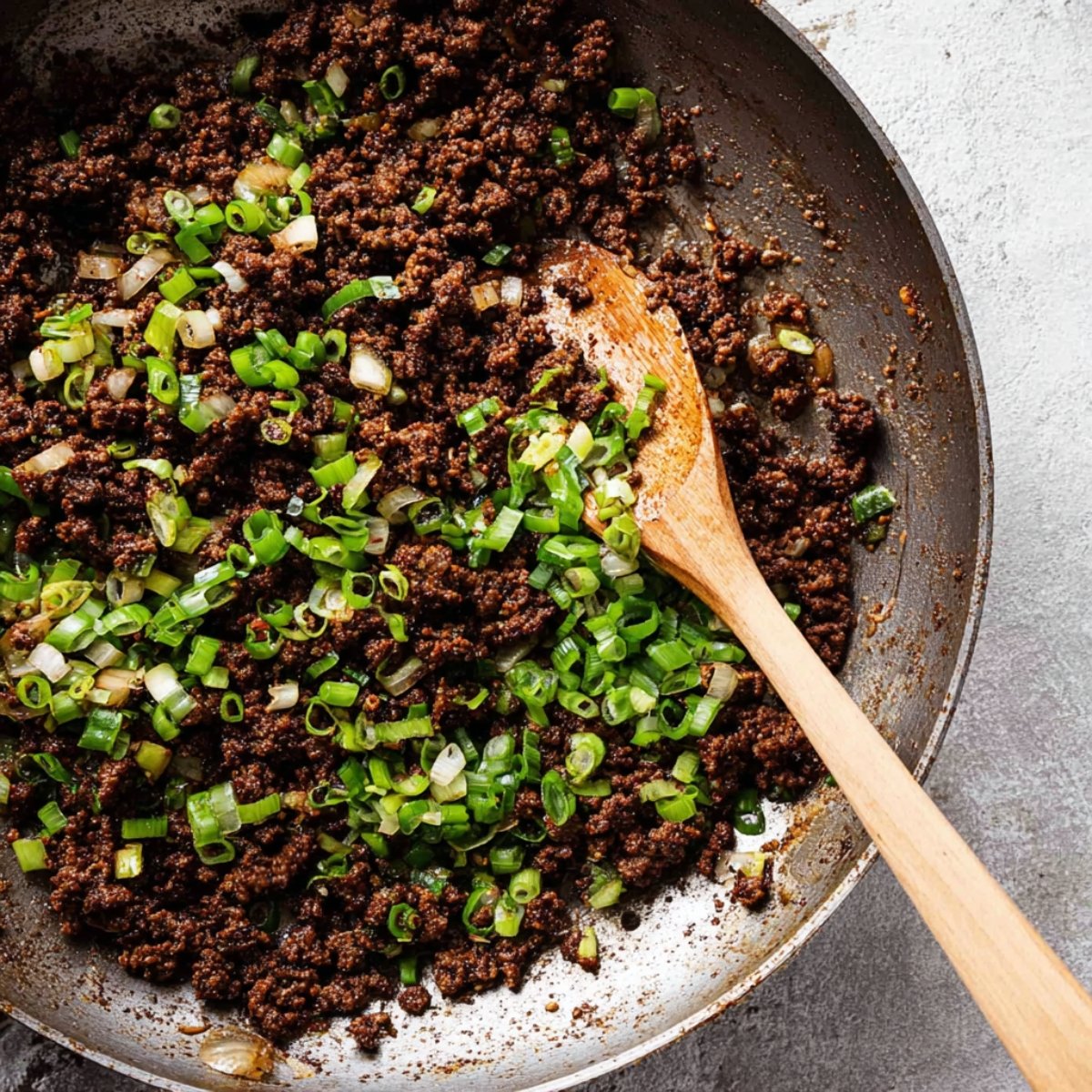 Ground beef cooked in a pan with green onions added on top, mixed together with a wooden spoon.