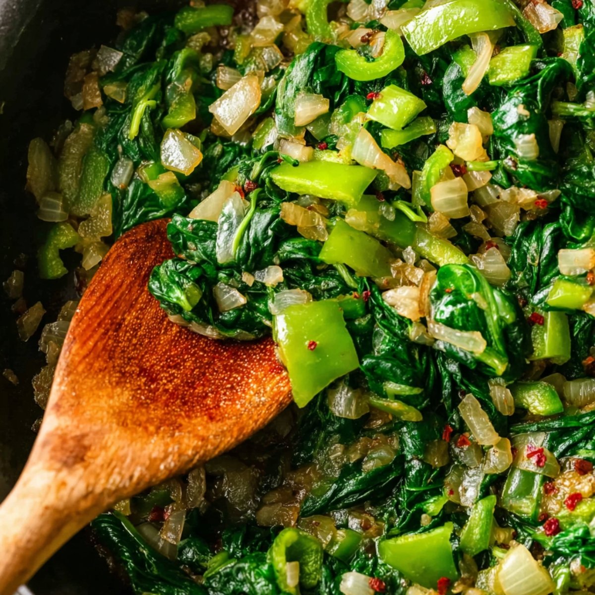 Close-up of sautéed spinach, onions, and green peppers in a skillet. The vegetables are cooked to perfection, with a wooden spoon stirring the mix, creating a vibrant and healthy dish.
