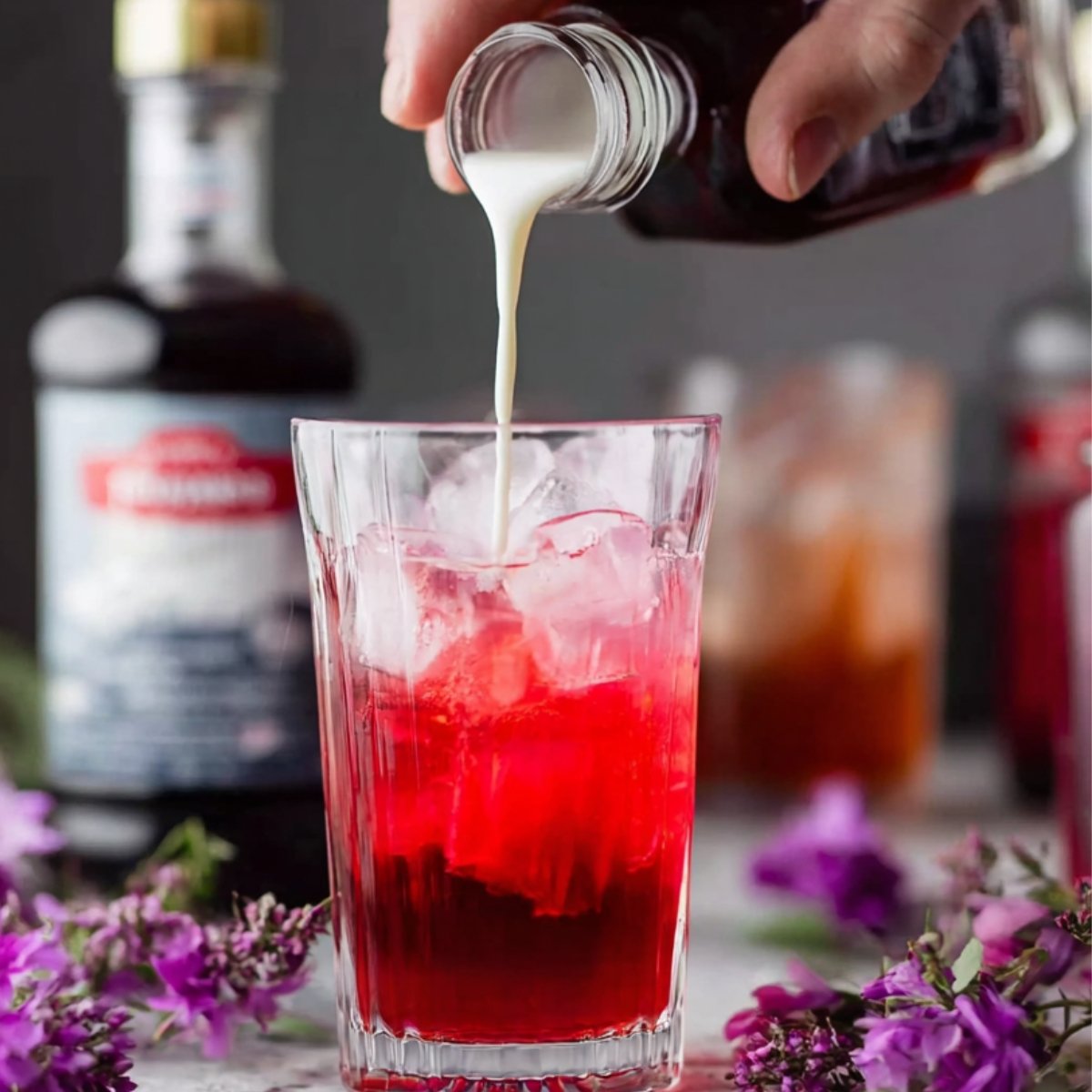 A close-up shot of a hand pouring cream into a glass filled with ice cubes and bright red soda. The image features vibrant purple flowers in the foreground and bottles of syrup in the background, enhancing the playful and colorful aesthetic.