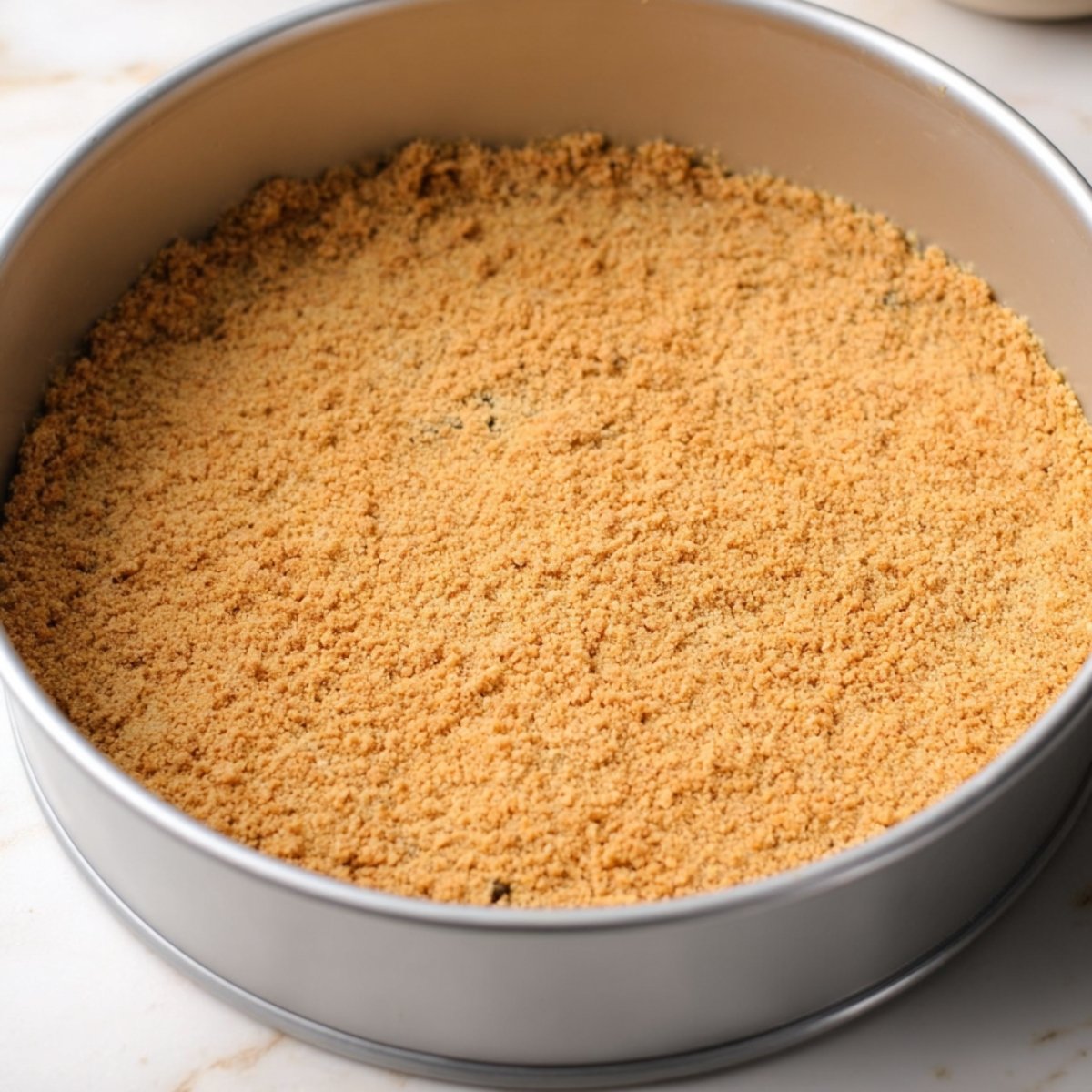 A round springform pan filled with an evenly pressed golden graham cracker crust, ready to be baked.