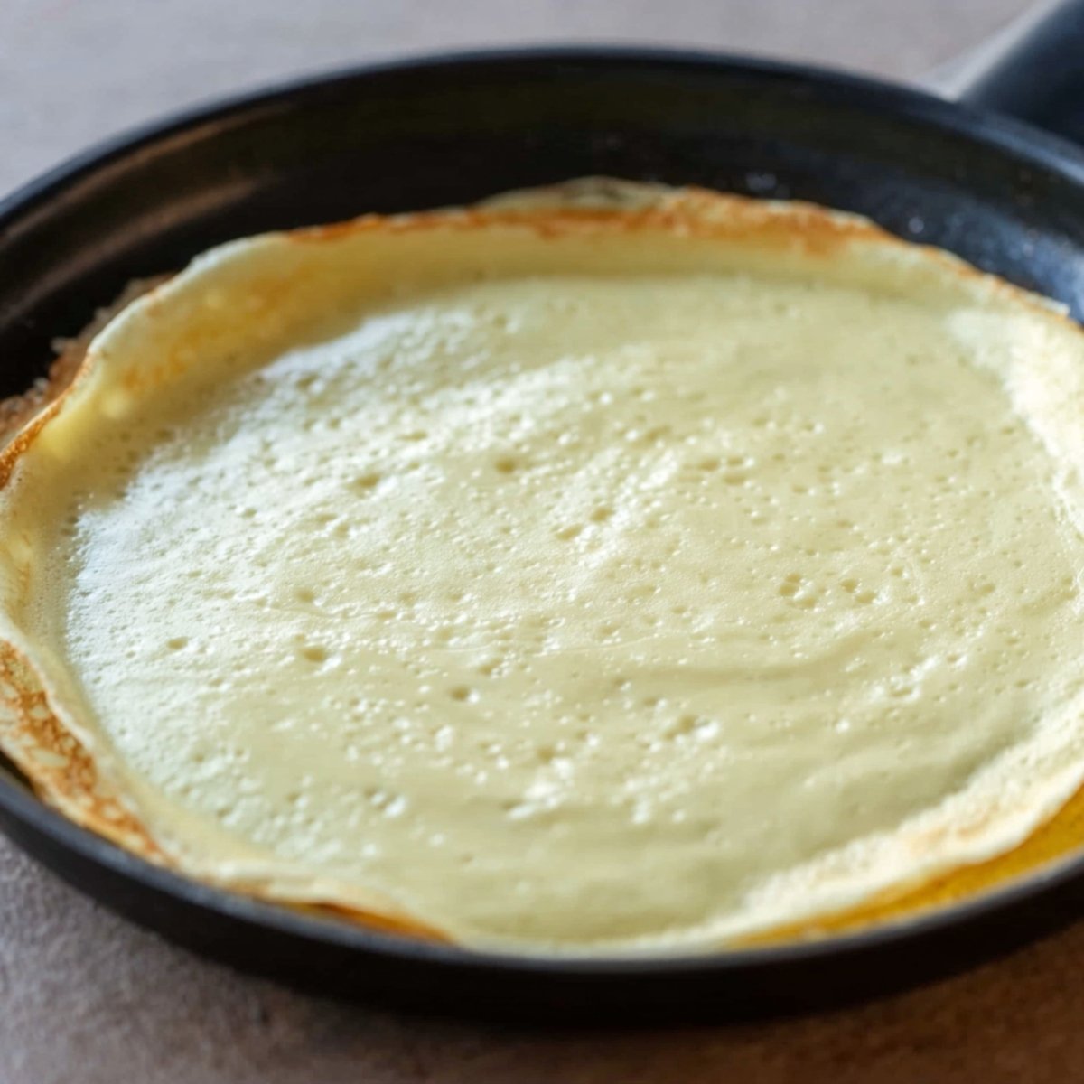 Close up of a crepe cooking in a skillet, with a smooth, golden batter spread evenly across the pan. The crepe is starting to form bubbles and edges, indicating it’s ready to flip.