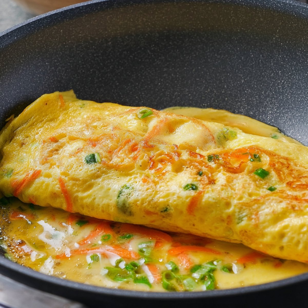 Close up of a Korean rolled omelette (Gyeran Mari) in a frying pan, with the egg mixture partially cooked and rolled, showcasing a bright yellow color with green onions and carrots.