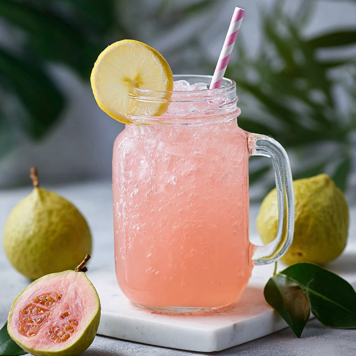 A mason jar glass filled with pink iced Guava White Tea Lemonade drink, garnished with a lemon slice and striped straw, surrounded by whole and halved guavas on a marble surface.