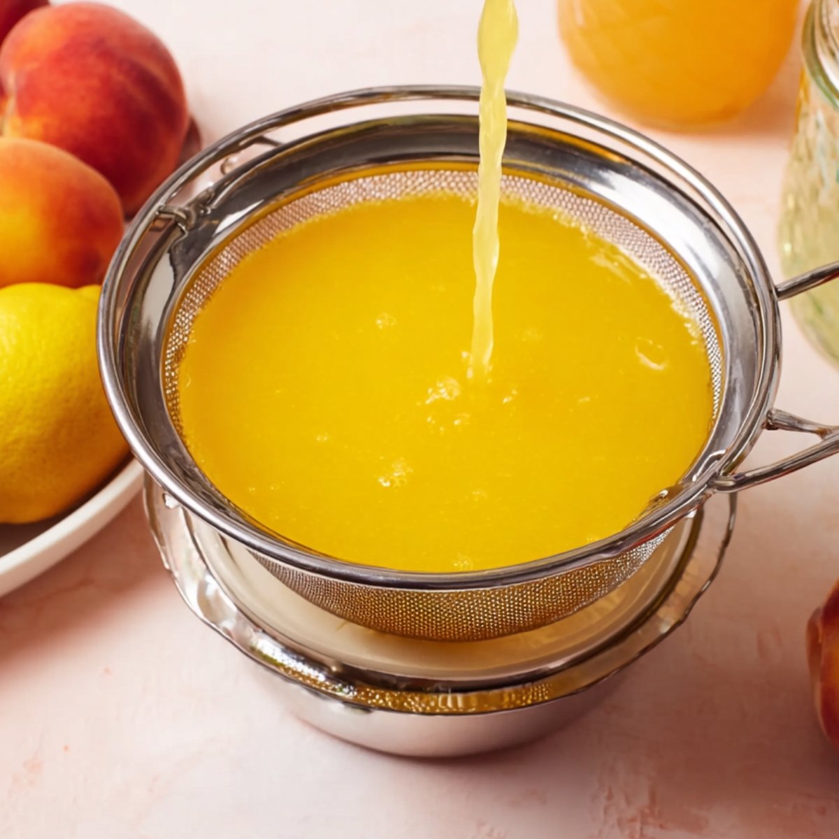 Close up of freshly strained peach and lemon juice flowing through a fine mesh sieve into a bowl, with fresh peaches and lemons around the scene."