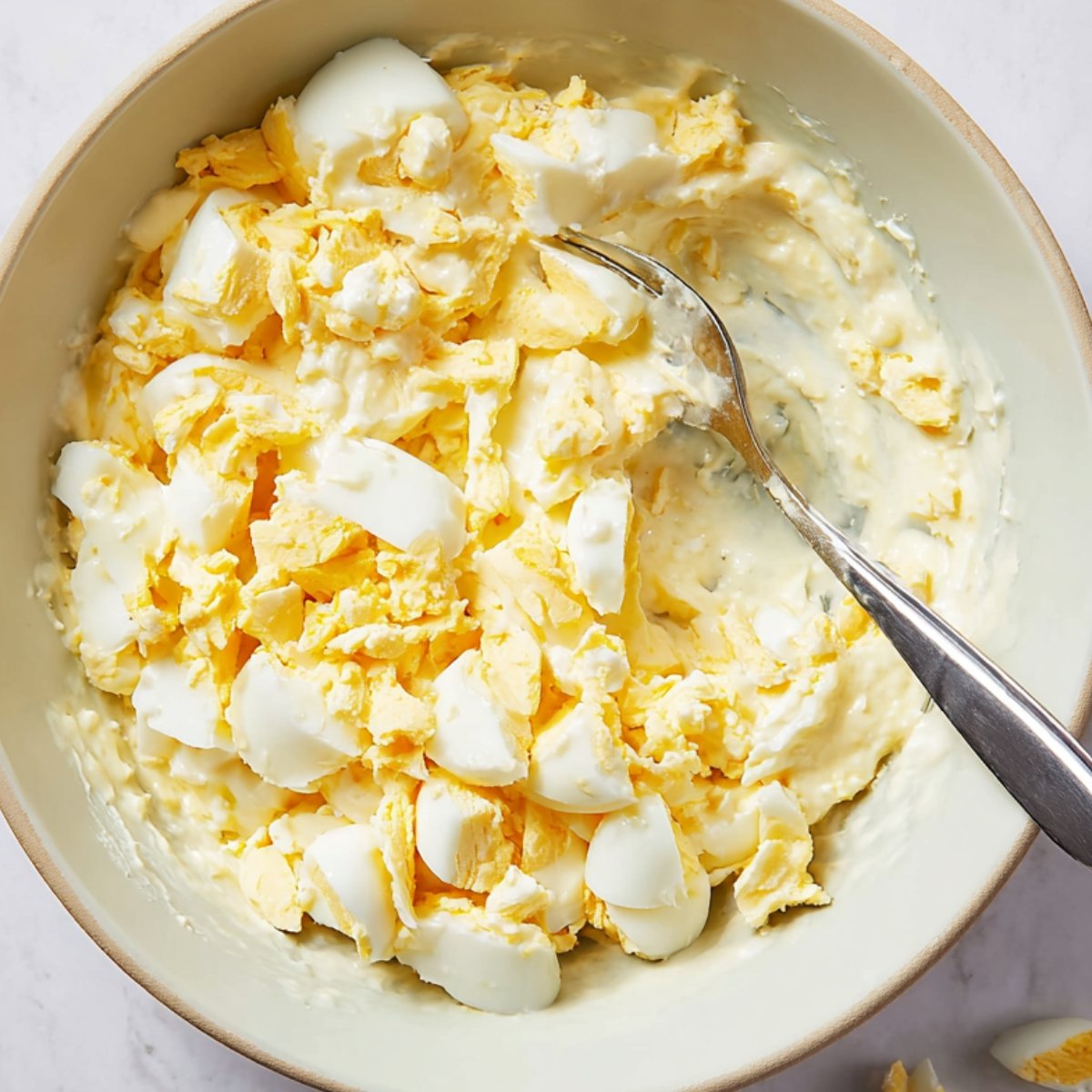 Close up of a bowl containing chopped boiled eggs mixed with creamy mayonnaise, a spoon gently stirred in the filling, ready to be spread on bread for sandwiches."