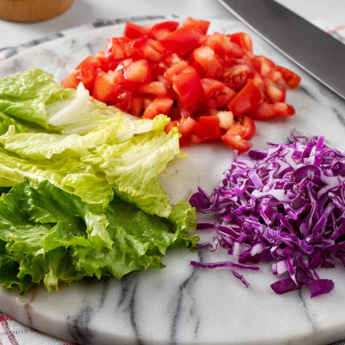 Chopped tomatoes, shredded purple cabbage, and romaine lettuce on a marble cutting board, with a knife beside the ingredients.