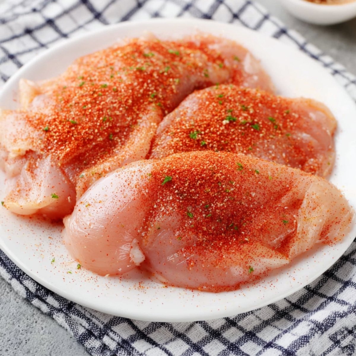 Raw chicken breasts sprinkled with paprika and seasoning, arranged on a white plate, resting on a checkered cloth.