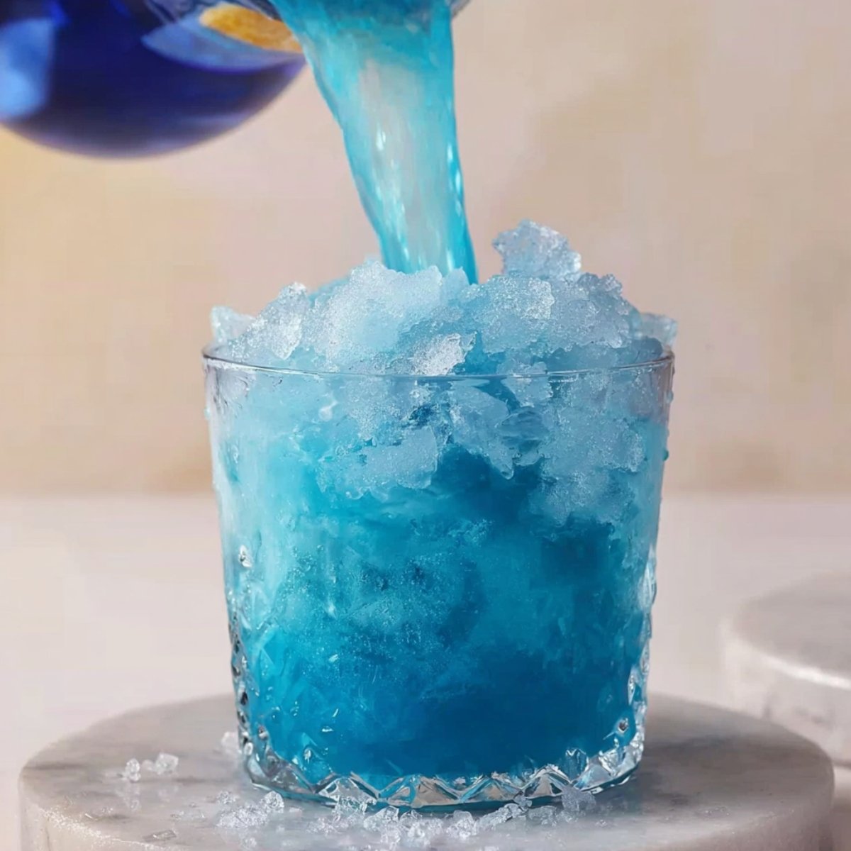 "Close up of a blue drink being poured over crushed ice in a clear glass, creating a refreshing, icy layer of blue. The glass sits on a marble surface, hinting at a tropical, cool beverage.