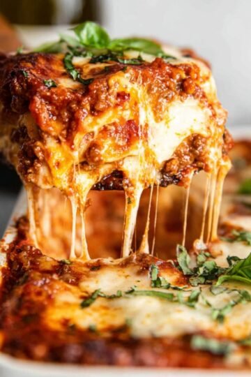 Close-up of a slice of homemade lasagna being lifted from a baking dish, with layers of rich meat sauce, melted cheese, and a sprinkle of fresh basil. The cheesy layers stretch invitingly.
