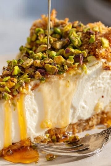 A close-up of a slice of Baklava Cheesecake topped with pistachios, drizzled with syrup. The slice has a rich creamy texture, with the crunchy pistachio topping adding a contrast.