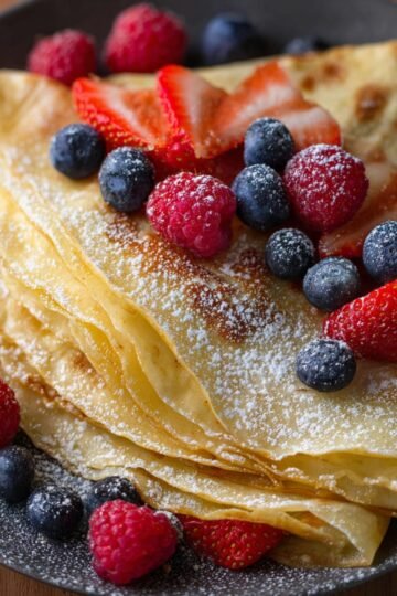 A stack of delicious Crepe Recipe garnished with fresh strawberries, raspberries, and blueberries, lightly dusted with powdered sugar, ready to be served as a sweet breakfast or dessert.
