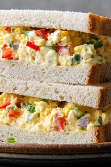 Close up of a stack of egg mayo sandwich, made with white bread and filled with a creamy, colorful mixture of chopped eggs, red bell pepper, and green onions, looking appetizing and fresh.