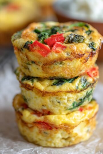Close-up of stacked, golden, Fluffy Cottage Cheese Egg Muffins with spinach and red pepper, resting on parchment paper, with a side of cottage cheese in the background.