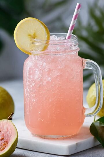 A mason jar glass filled with pink iced Guava White Tea Lemonade drink, garnished with a lemon slice and striped straw, surrounded by whole and halved guavas on a marble surface.