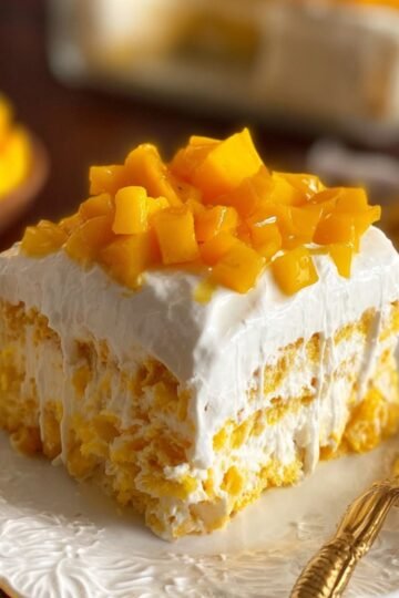 A slice of Mango Tres Leches Cake dessert layered with cream and cake, topped with diced mango, served on a white plate with a gold fork.