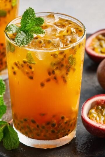 Close up of a glass of passion fruit iced tea with ice cubes, fresh mint leaves, and floating passion fruit pulp, served with passion fruit halves in the background.