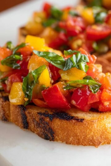 Close up of a vibrant, freshly made Tomato Bruschetta topped with diced tomatoes, yellow peppers, and fresh basil, served on toasted bread with a golden, crispy finish.